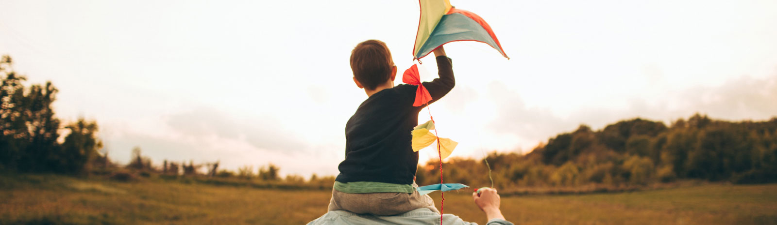 young boy riding on dad's shoulders and holding a multi-colored kite